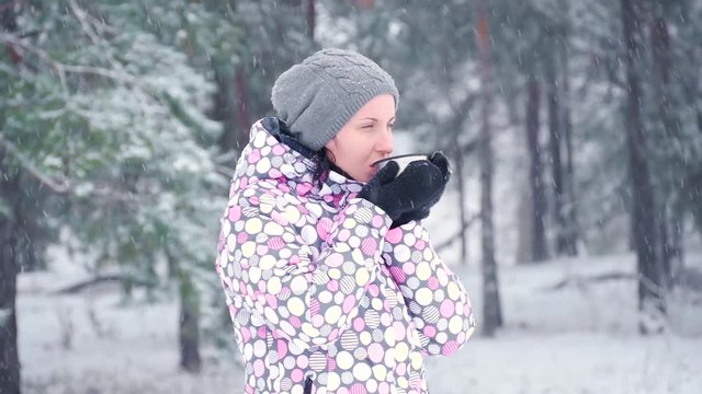 A freezing tourist woman drinks hot tea from a mug against the backdrop of a winter forest or park on a snowy day. Slow motion