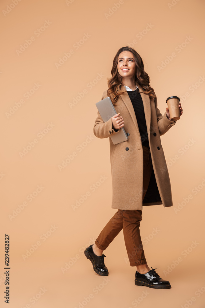 Poster image of beautiful woman 20s wearing coat holding cell phone and takeaway coffee while walking, isol