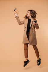 Image of elegant woman 20s wearing coat holding cell phone and takeaway coffee while walking, isolated over beige background