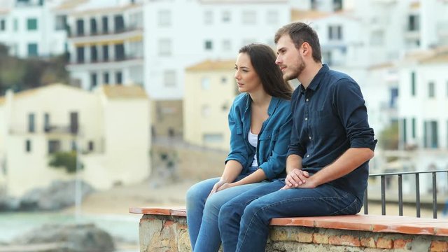 Serious couple of friends talking looking away sititng on a ledge on vacation in a coast town