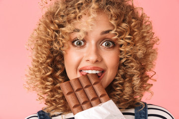 Photo of joyous curly woman 20s eating chocolate bar while standing, isolated over pink background