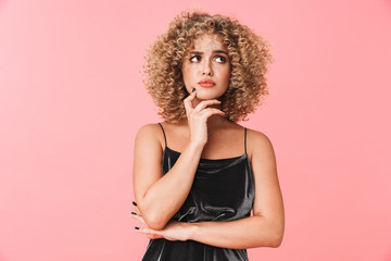 Portrait of serious curly woman 20s wearing dress thinking and touching chin, isolated over pink background