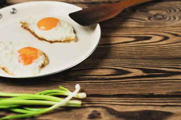 Two fried eggs in metal beige pan and dirty spatula and green fresh onion on old wooden brown table. Copy space