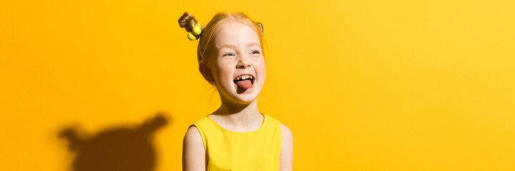 Girl with red hair on a yellow background. The girl shows her tongue. - obrazy, fototapety, plakaty