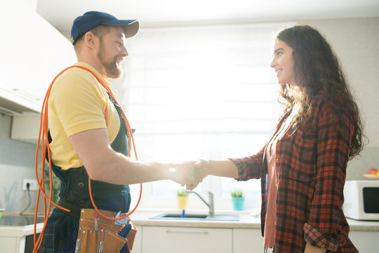 Smiling young lady with curly hair shaking hand of content repairman with wire on shoulder while thanking him for good job