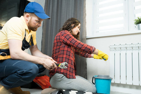 Serious attractive young lady in casual shirt and rubber gloves squeezing cloth while mopping floor from radiator water, repairman in cap sitting near her and waiting when he will start working