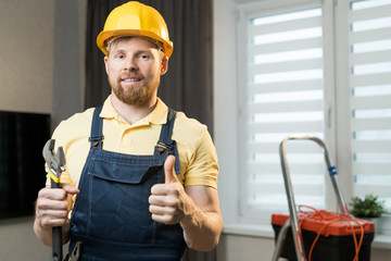 Smiling satisfied young bearded repairman in hardhat standing in modern room and holding wrench while showing thumb-up and looking at camera