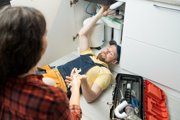 Smiling handsome young plumber in cap lying on floor in kitchen and fixing sink pipe while home owner giving wrench to him - obrazy, fototapety, plakaty