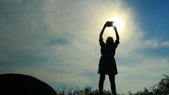 Silhouette of young woman in dress standing on the rock and takes a picture with mobile phone. slow motion. 1920x1080