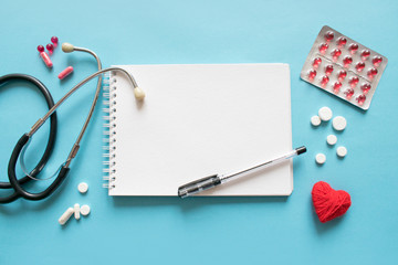 Medical mockup with stethoscope, pills drugs, red heart, white notepad on blue background. Doctor work place flat lay, top view with copy space