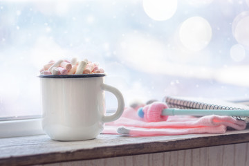 Obraz na płótnie Canvas Winter cozy concept. Coffee with marshmallows, pen with cap and pink gloves near window sill with the snow-covered window bokeh. Warm weekend in cold weather