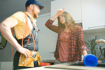 Exhausted young woman with curly hair wiping forehead and holding plunger while talking to young...