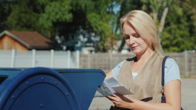 A woman looks at the envelopes with letters, then throws them into the blue mailbox on the street