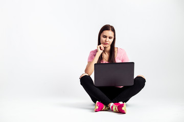 Pretty girl dressed in pink t-shirt and jeans is sitting on the floor with laptop on the white background in the studio