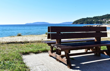 Bench in a beach promenade with grass and vegetation in sand. Blue sea, clear sky, sunny day. Galicia, Spain.