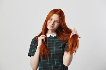 Beautiful redhead long-haired girl in a green checkered dress holds her hand on her hairs on the white background in the studio