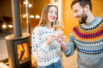 Young couple in sweaters celebrating winter holidays standing with sparkling wine in the modern house with fire place