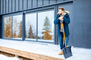 Portrait of a man in winter clothes standing with snow shovel on the terrace of the building in the...