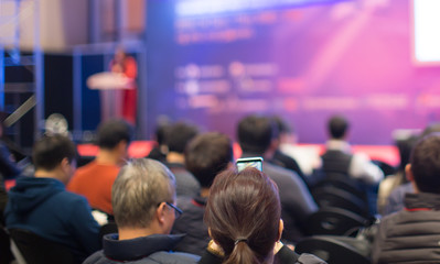 "Audience Watching a Presentation. Business. Female Presenter on Stage Giving Talk to Crowd of People. Woman Speaker at  Investor Pitch Conference.  Defocused Blurred Presenter During Conference."
