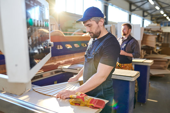 Serious concentrated manual worker with beard wearing cap and denim overall standing at workbench of industrial machine and choosing wooden pattern