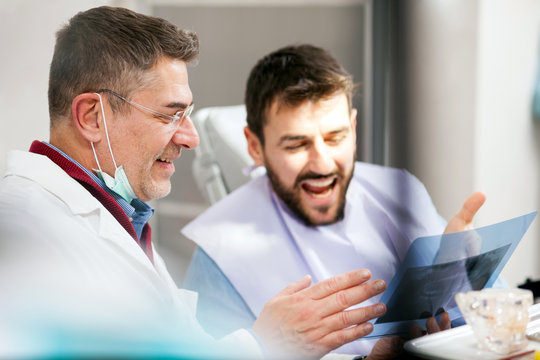Smiling mature male dentist and young patient looking at teeth x-ray image after successful medical intervention. Health care and medicine concept