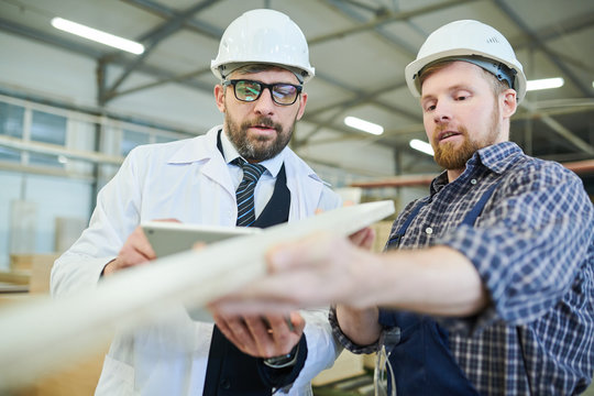 Content bearded worker in white hardhat showing wooden plank to middle-aged inspector in glasses while they checking surface of manufactured product