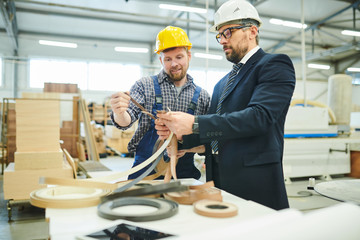 Serious modern investor in hardhat standing at table and analyzing quality of manufactured products...