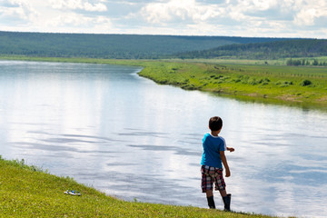 Fototapeta na wymiar Yakut boy in rubber boots on the precipice above the river at summer sunny day. Back view. Empty space for text