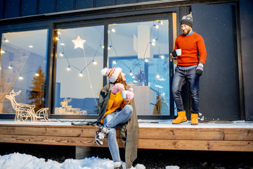 Young couple warming up with plaid and hot drinks sitting on the terrace of the modern house in the mountains durnig the winter holidays