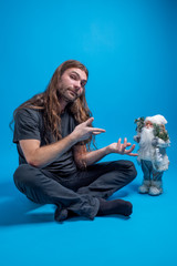 Naklejka premium Long-haired man pointing to a toy Santa while sitting down. Studio portrait shot against blue background