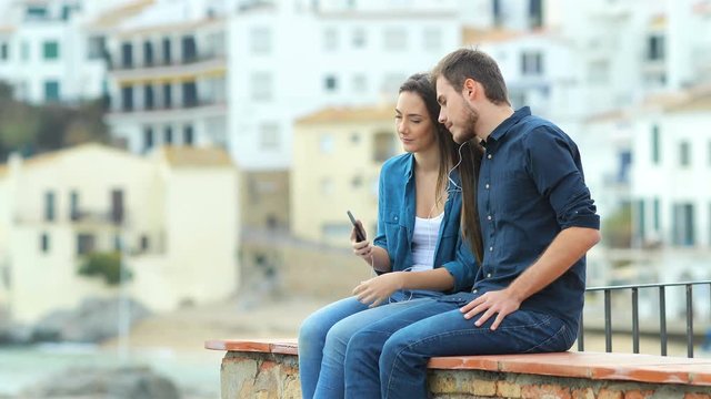 Serious couple sharing music and breathing outdoors sitting on a ledge on vacation