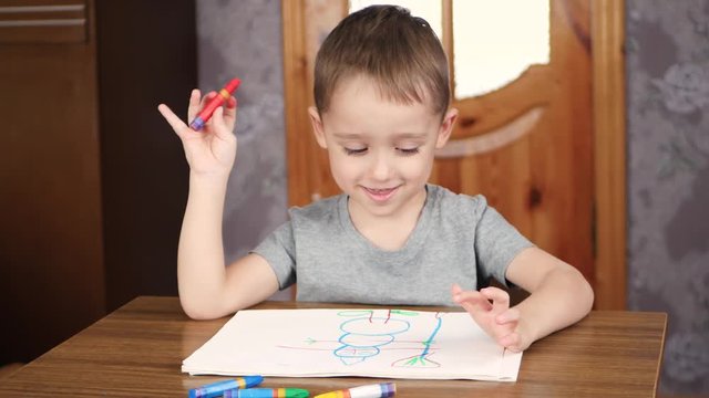 A happy smiling cute child draws with bright crayons or a pencil while sitting at a table at home. The development of children of preschool age, creativity.