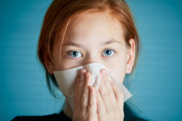 Sick girl with blue beautiful eyes holding white napkin at nose for blowing out.