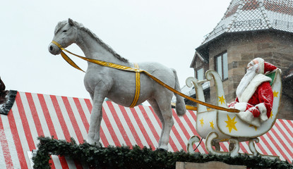 Weihnachtsmann auf weißem Schlitten mit goldenen Sternen wird von einem Pferd gezogen. Auf einem Dach von einem weiß-roten Stand auf dem Nürnberger Christkindlesmarkt.