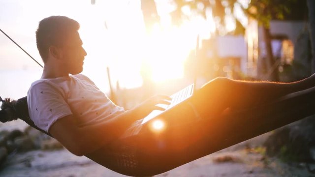 A young, handsome man working on a laptop lying in a hammock at sunset. Freelancer works on travel.
