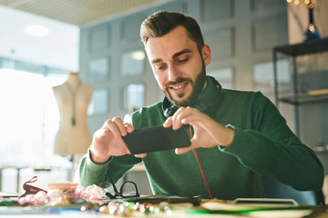 Portrait of handsome young man taking photo via smartphone while sitting at desk in sunlit office and smiling, copy space