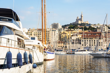 Luxury yachts and sailboats moored in the marina of the Old Port of Marseille, France, with Notre-Dame de la Garde basilica on top of the hill at sunset.