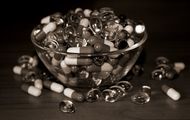 Pills in glass bowl on wood background. Scattering of tablets of different shapes and colors. Healthy lifestyle, possibility of addiction to medication and excess dosage. Toning in shades of brown.
