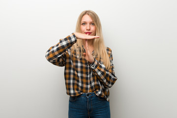 Blonde young girl over white wall making stop gesture with her hand to stop an act