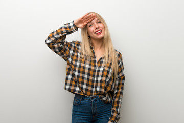 Blonde young girl over white wall saluting with hand