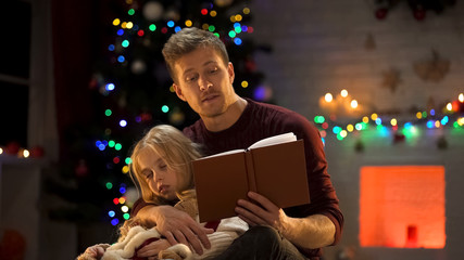 Attentive dad reading Christmas fairy-tale for sleepy girl near decorated tree