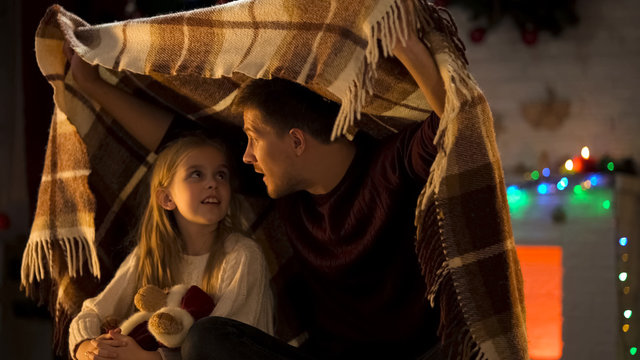 Father telling breathtaking X-mas story for little girl sitting under cozy plaid