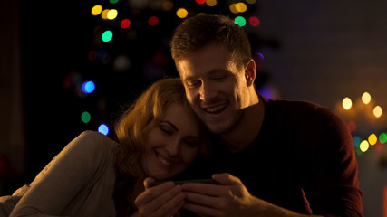 Smiling couple watching movie on smartphone under Christmas tree, fun mood