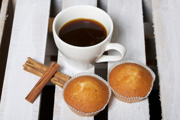 A cup of freshly brewed coffee and muffins. Nearby are cinnamon sticks. Located on a wooden box.