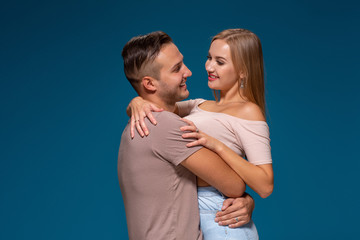 Young couple is hugging on blue background in studio. They wear T-shirts, jeans and smile.