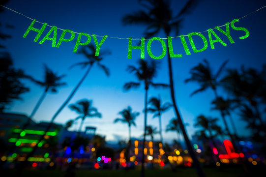 Happy Holidays message hanging in glittery party banner above tropical palm trees of the glowing neon lights of the skyline of Miami Beach, Florida, USA