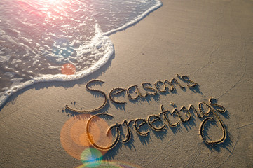 Season's Greetings message handwritten in smooth sand with an oncoming wave in the lens flare of the tropical sun