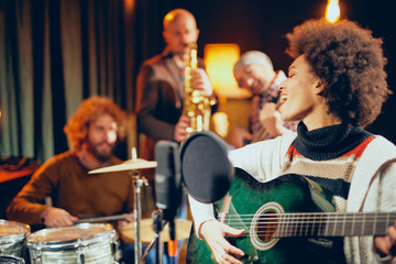 Mixed race woman singing and playing guitar while sitting on chair with legs crossed. In background drummer, saxophonist and bass guitarist. - obrazy, fototapety, plakaty