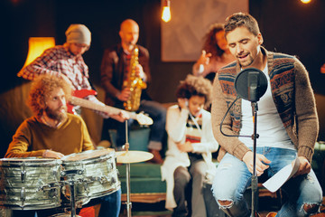 Man singing and sitting on chair while his band playing instruments in background. Home studio interior.