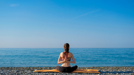 Young slim woman in tight sportswear sitting on orange yoga mat and practicing outdoors at pebble beach by the sea. Ypga at nature concept. Girl standing with prayer hands